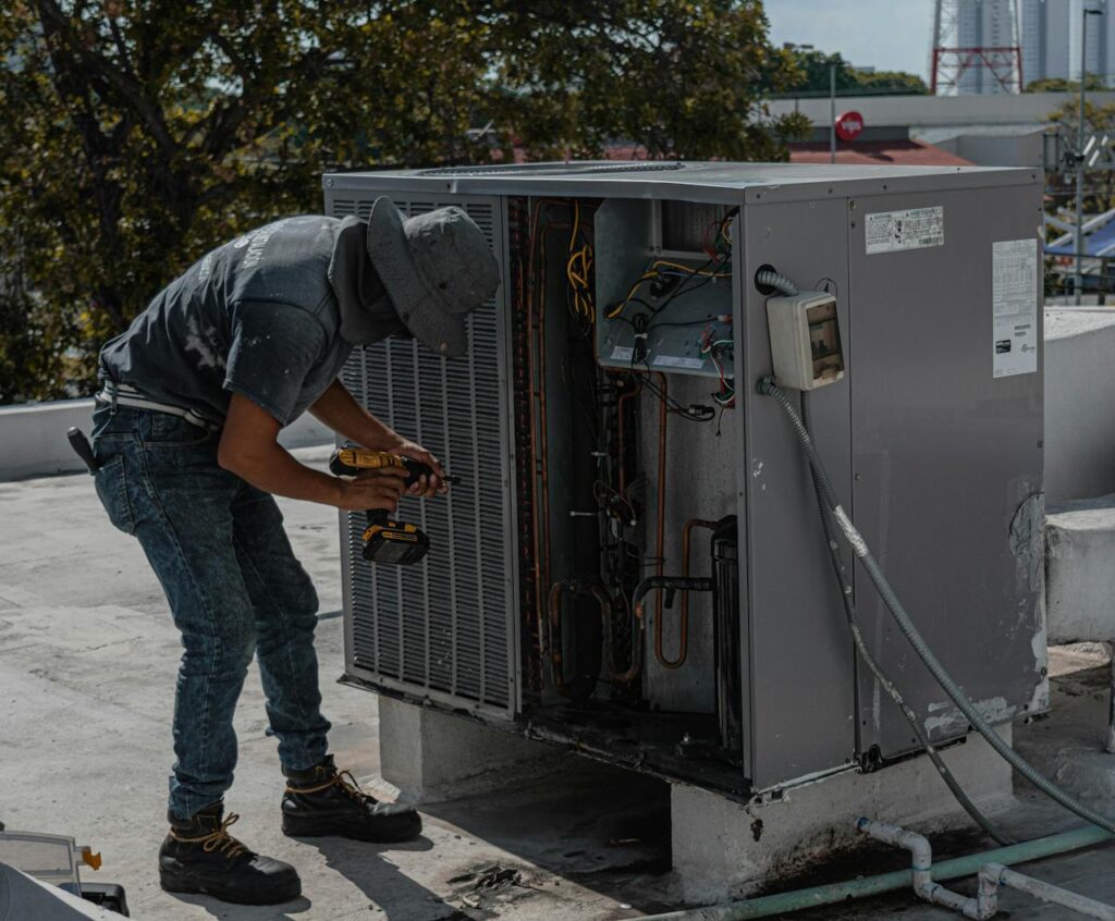 A worker in a bucket hat repairs an outdoor air conditioning unit on a rooftop.